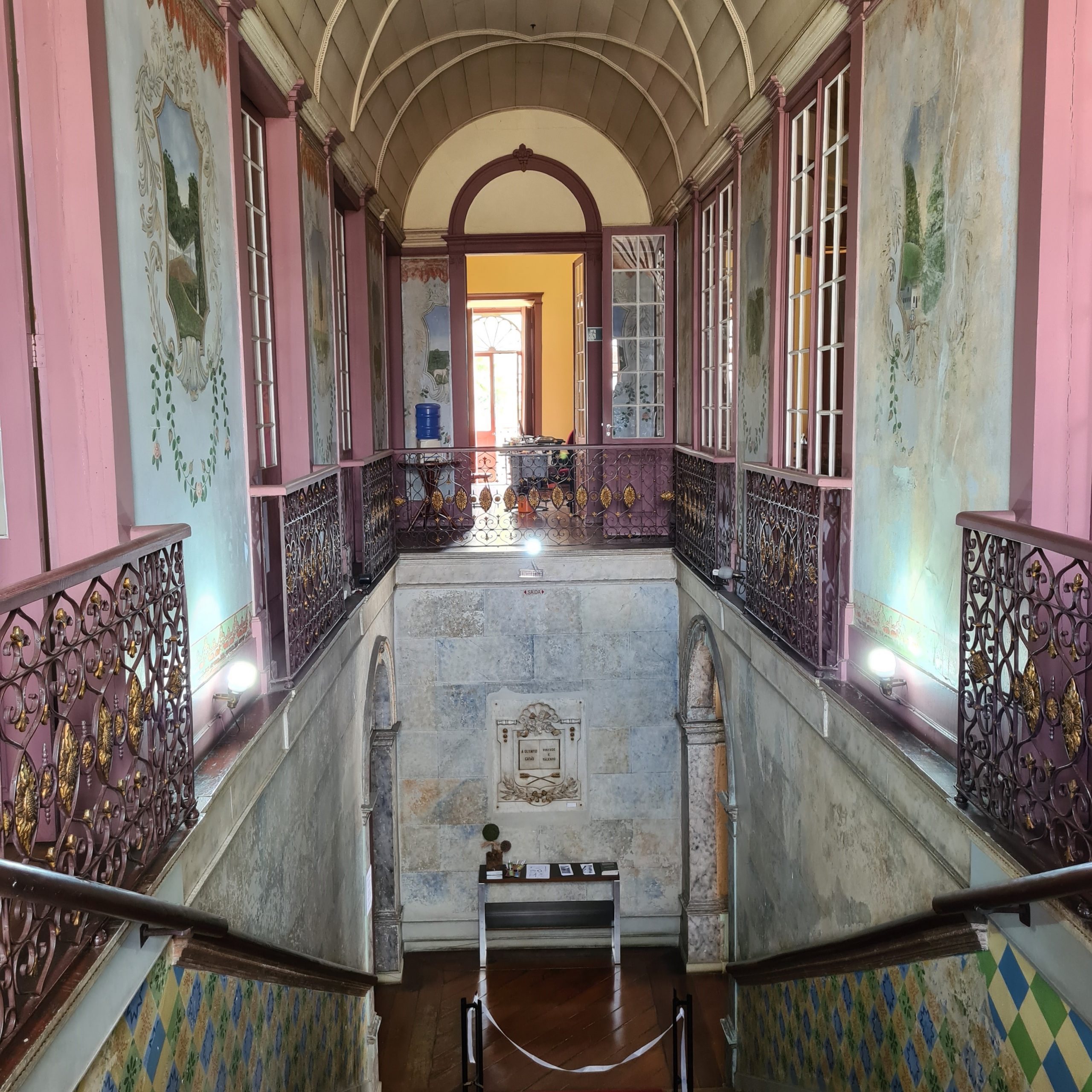 Imagem representa o acesso a uma escadaria com paredes ornamentadas, com sacadas de metal ornamentado dando acesso pelo piso superior.