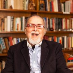 Imagem de Jorge Cunha Lima. Ele é um homem branco, cabelos e barbas grisalhos, usando um óculos de aros escuros, paletó preto e camisa listrada. Sorri para a foto à frente de uma estante de livros.